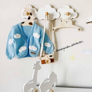 Puede incluir: Un cárdigan azul claro con aplicaciones de nubes blancas cuelga de una percha de madera con forma de jirafa. También se exhiben una cámara de madera con una correa blanca y una percha con forma de oso. Dos juguetes de animales de madera.