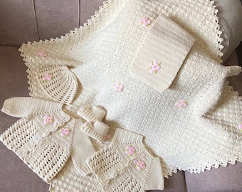 May include: A set of crocheted baby clothes in cream with pink flower accents. The set includes a cardigan, a vest, a pair of booties, and a blanket.