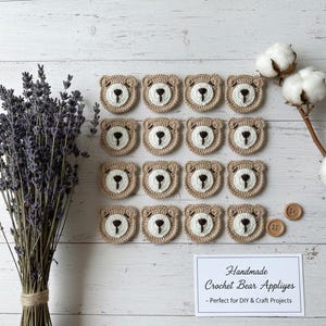 Könnte beinhalten: Handgefertigte Häkel-Bärenapplikationen in Beige und Weiß, angeordnet auf einer weißen Holzoberfläche. Die Applikationen zeigen ein Bärengesicht-Design. Das Bild enthält auch getrockneten Lavendel, Baumwollzweige und Holzknöpfe.