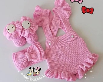 May include: A pink knitted baby outfit set with a headband, booties, and a sleeveless romper with ruffles. The romper has straps and a button closure. The headband has a bow.
