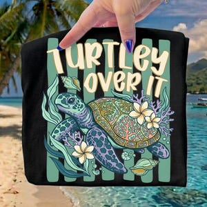 May include: Black tote bag featuring a colorful sea turtle design with the text "TURTLEY OVER IT". The design includes a sea turtle, flowers, and seashells. The background shows a tropical beach scene.