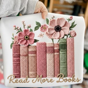 May include: White decorative pillow featuring a floral and book design. The design includes pink flowers, green leaves, and a row of books in shades of pink and green. The text "Read More Books" is written in a cursive font.