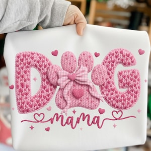 May include: White sweatshirt with pink embroidered text and design. The word "DOG" is spelled out in a knitted font, with a paw print in the center. The word "mama" is embroidered below. Pink hearts and stars are scattered around the design.