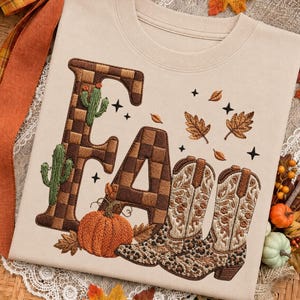 May include: A beige crewneck sweatshirt featuring an embroidered design with the word FALL. The letters F and A are filled with a brown checkered pattern, while the L is formed by two cowboy boots with floral and leopard print details.