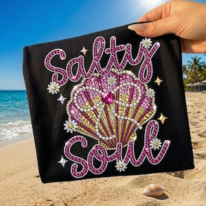 Puede incluir: Bolso tote negro con la frase "Salty Soul" en rosa con pedrería. El diseño presenta una concha con detalles rosas, amarillos y blancos, además de estrellas y flores.