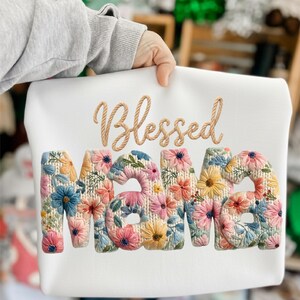 Könnte beinhalten: Weißes Sweatshirt mit dem gestickten Schriftzug "Blessed Mama". Das Wort "Mama" ist mit farbenfrohen Blumenstickereien in Rosa, Blau, Gelb und Orange gefüllt. Ein komfortables und stilvolles Design.