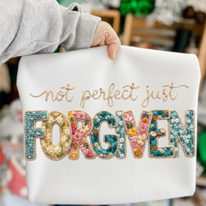 May include: White sweatshirt with the embroidered text "not perfect just FORGIVEN". The word "FORGIVEN" is in large, colorful floral letters. The sweatshirt is held by a person.