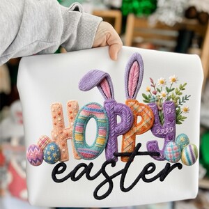 Puede incluir: Cojín decorativo blanco con temática de Pascua con las palabras "HOPE Easter" en un diseño colorido y texturizado. La "O" en "HOPE" tiene un patrón de rayas. También se presentan orejas de conejo y huevos de Pascua.