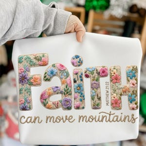 May include: White sweatshirt with the word "Faith" embroidered in colorful floral letters. The text "can move mountains" is embroidered below. The sweatshirt is held by a person.