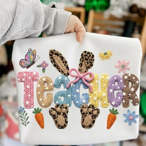 May include: White sweatshirt featuring the word "Teacher" in colourful, textured letters. The design includes a butterfly, bunny ears, paw prints, carrots, and flowers, creating a festive, spring-themed look.