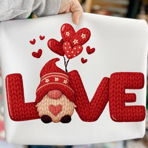 Op de afbeelding: Wit kussen met een rood gebreid "LOVE" ontwerp. De "O" is vervangen door een kabouter met een rode hoed en hartvormige ballonnen. Kleine rode harten zijn verspreid rond de ballonnen.