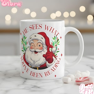 May include: White ceramic mug featuring a cartoon Santa Claus winking and pointing. The mug has the text "HE SEES WHAT YOU'VE BEEN READING" in red. The Santa illustration is surrounded by a green wreath and red stars.