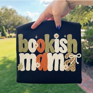 May include: Black tote bag with the embroidered text "bookish mama" in a mix of colors and textures. The letters are in a knitted style, with floral and leaf accents. The bag is held up against a background of green foliage.