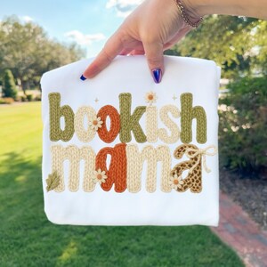 May include: White t-shirt with the text "bookish mama" in a knitted style. The letters are in shades of green, orange, and cream, with daisy and leaf accents. The shirt is held up by a person.