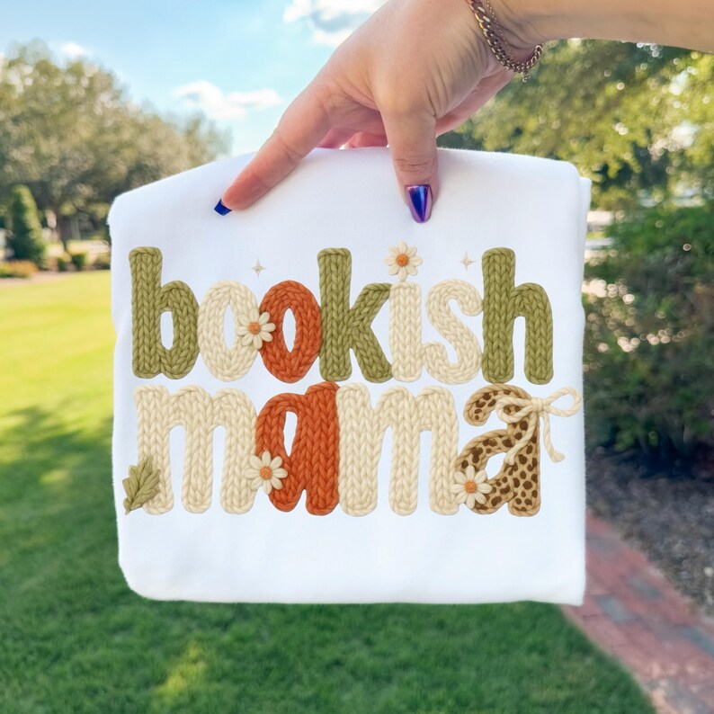 May include: White t-shirt with the text "bookish mama" in a knitted style. The letters are in shades of green, orange, and cream, with daisy and leaf accents. The shirt is held up by a person.
