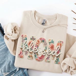 May include: Cream-colored sweatshirt with the word "MOM" and the word "blessed" embroidered in a floral design. The floral design includes pink, orange, and green flowers. A small butterfly is embroidered on the letter "O". A silver necklace with a pendant is draped on the sweatshirt.