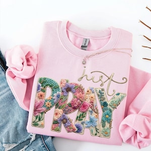 May include: A pale pink sweatshirt featuring the words "just PRAY" in a floral design. The letters are filled with colourful flowers and a knitted texture. A delicate necklace is draped across the sweatshirt.