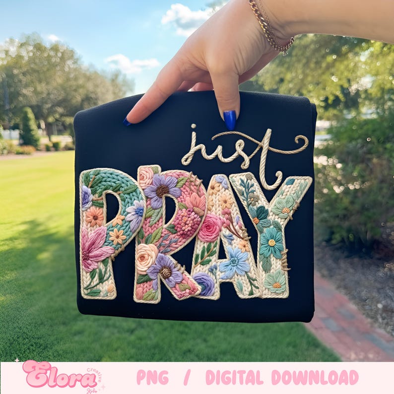 May include: A black square design featuring the embroidered words "just PRAY". The letters are filled with colourful flowers in shades of pink, blue, and purple. The design is held by a hand with blue nail polish.