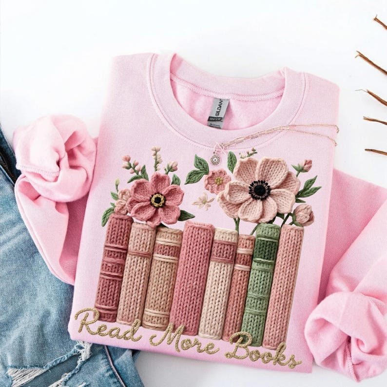 May include: A light pink sweatshirt with an embroidered design featuring a stack of books and flowers. The words "Read More Books" are embroidered in gold. A delicate necklace with a small pendant rests on the sweatshirt.