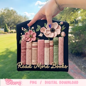 May include: A black square with embroidered books and flowers. The books are in shades of pink, cream, and green. The phrase "Read More Books" is written in gold script.