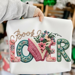 May include: White sweatshirt featuring the embroidered words "Book Lover". The design incorporates floral and knitted elements in pink, green, and beige. The sweatshirt is held by a person.