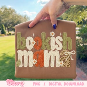 May include: A brown sweatshirt with the words "bookish mama" in a knitted style. The letters are in white, green, and orange with daisy accents. The bottom right of the sweatshirt has a leopard print bow.