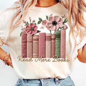 May include: Cream-colored t-shirt featuring a floral and book design. The design includes pink flowers, green leaves, and a row of books in shades of pink and green. The text "Read More Books" is in a gold script.