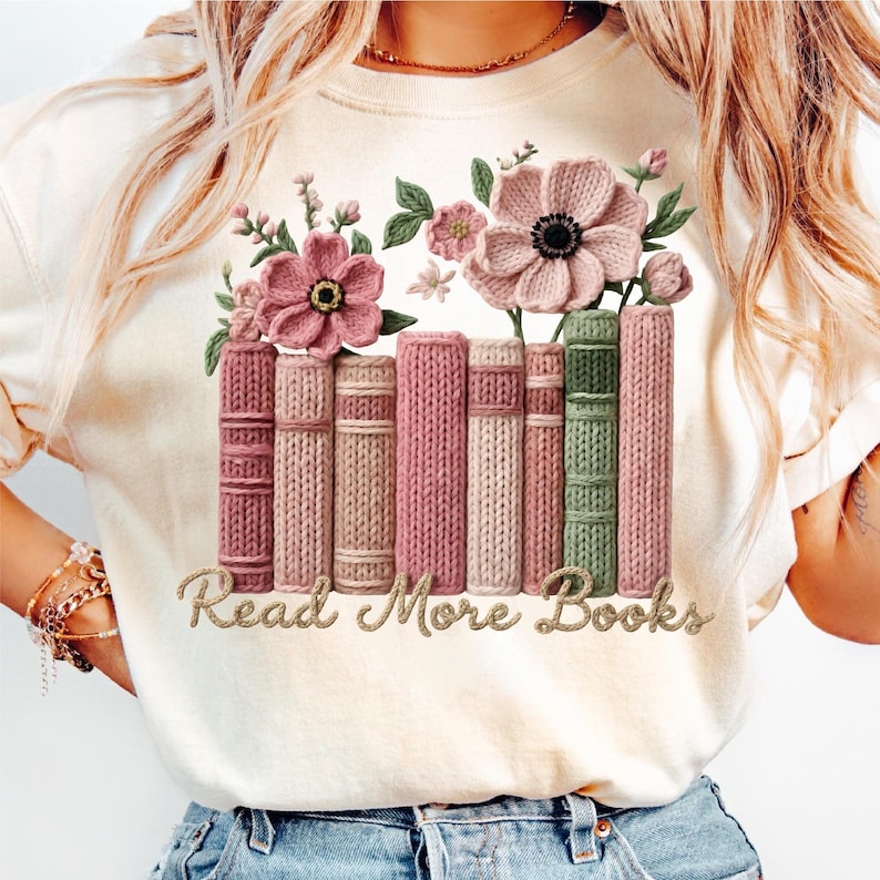 May include: Cream-colored t-shirt featuring a floral and book design. The design includes pink flowers, green leaves, and a row of books in shades of pink and green. The text "Read More Books" is in a gold script.