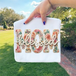 May include: White t-shirt with the word "MOM" in floral embroidery, and the word "blessed" in a cream-colored script. A butterfly is embroidered on the letter "O". The shirt is held up by a hand.