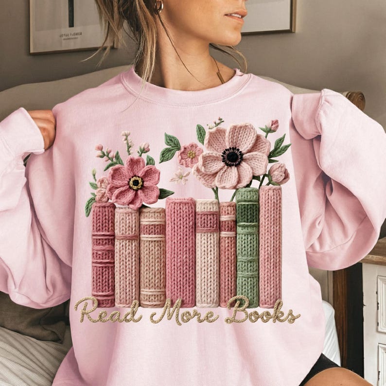 May include: A light pink sweatshirt featuring a floral and book design. The design includes pink flowers, green leaves, and a stack of books in various shades of pink and green. The text "Read More Books" is embroidered in gold.