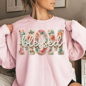 May include: A light pink sweatshirt with the words "MOM blessed" in a floral design. The letters are filled with pink, orange, and green flowers, and a small butterfly is visible. The sweatshirt is a soft, casual style.