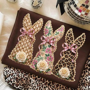 May include: A brown decorative textile featuring three bunny silhouettes with ornate designs. Each bunny has a different pattern: gold and cream, floral, and leopard print. They are adorned with pink bows and rose accents.