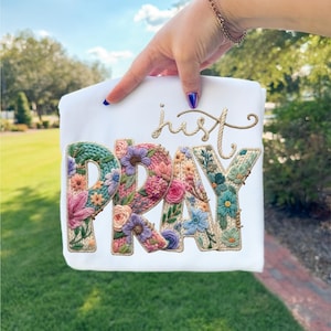 May include: White t-shirt featuring the embroidered words "just PRAY". The letters are filled with colourful floral designs in shades of pink, blue, green, and purple. The word "just" is written in a cursive font.