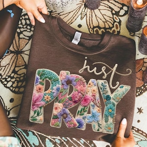 May include: A brown sweatshirt featuring the embroidered words "just PRAY". The letters are filled with colourful flowers in shades of pink, blue, and green. The sweatshirt is held by two hands.