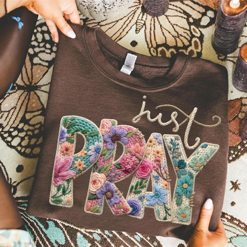 May include: A brown sweatshirt featuring the embroidered words "just PRAY". The letters are filled with colourful flowers in shades of pink, blue, and green. The sweatshirt is held by two hands.