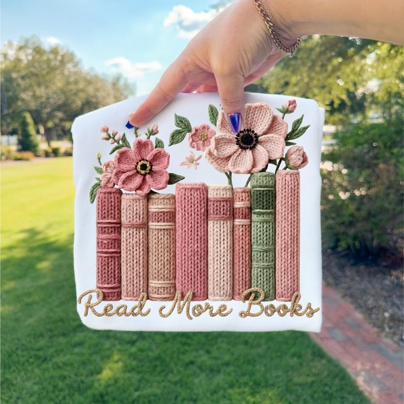 May include: White t-shirt featuring a floral and book design. The design includes pink flowers and a row of books in shades of pink and green. The text "Read More Books" is written in gold script.