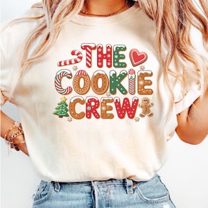 May include: A cream-coloured t-shirt featuring the text "THE COOKIE CREW" in a gingerbread-style font. The design incorporates candy canes, gingerbread men, and Christmas tree graphics, ideal for the festive season.
