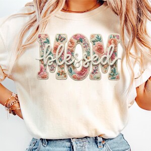 May include: Cream-colored t-shirt featuring the word "MOM" in large, floral-filled letters, with the word "Blessed" in a cursive font overlaying the letters. A small butterfly is visible on the letter "O".
