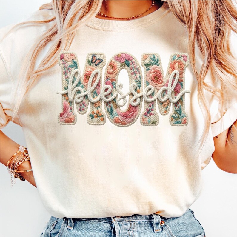 May include: Cream-colored t-shirt featuring the word "MOM" in large, floral-filled letters, with the word "Blessed" in a cursive font overlaying the letters. A small butterfly is visible on the letter "O".