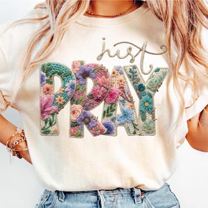 May include: A cream-coloured t-shirt with the words "just PRAY" in a floral design. The letters are filled with colourful flowers and a knitted texture, with the word "just" in a rope-like font.