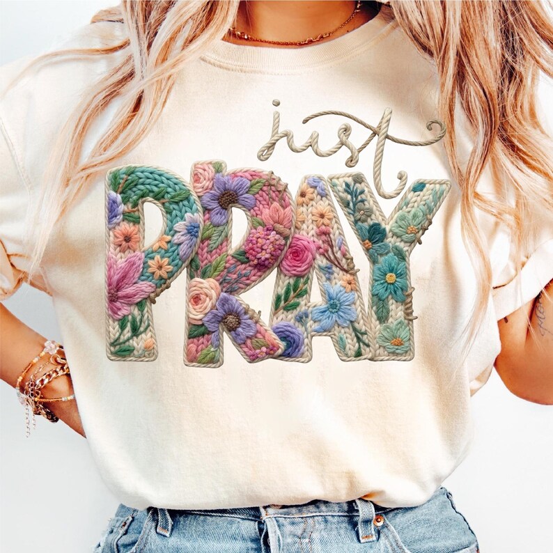 May include: A cream-coloured t-shirt with the words "just PRAY" in a floral design. The letters are filled with colourful flowers and a knitted texture, with the word "just" in a rope-like font.