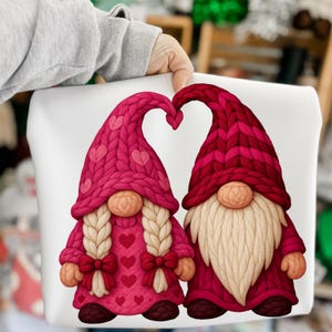Könnte beinhalten: Ein weißes Kissen mit zwei Zwergen mit gestrickten Hüten, die ein Herz bilden. Die Zwerge haben lange Bärte und sind in Rot- und Rosatönen gekleidet, ideal für den Valentinstag oder ein romantisches Thema.