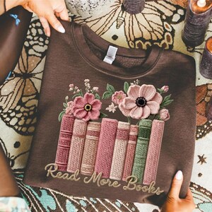 May include: A brown sweatshirt featuring an embroidered design of pink flowers and a stack of books in shades of pink and green. The text "Read More Books" is embroidered in a cursive font.