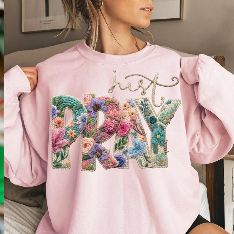 May include: A pale pink sweatshirt featuring the words "just PRAY" in a floral design. The word "PRAY" is filled with colourful flowers and leaves. The word "just" is written in a cursive font.