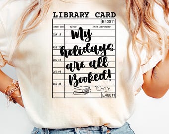 May include: Cream-colored t-shirt featuring a black graphic of a library card with the text "My holidays are all Booked!". The design includes a stack of books and glasses. The shirt is a casual wear item.