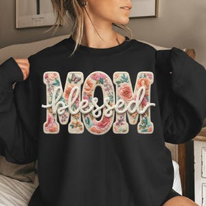 May include: Black sweatshirt with the words "MOM blessed" in a floral design. The letters are filled with pink, peach, and green flowers, and a small butterfly is visible. The word "blessed" is in a cursive font.