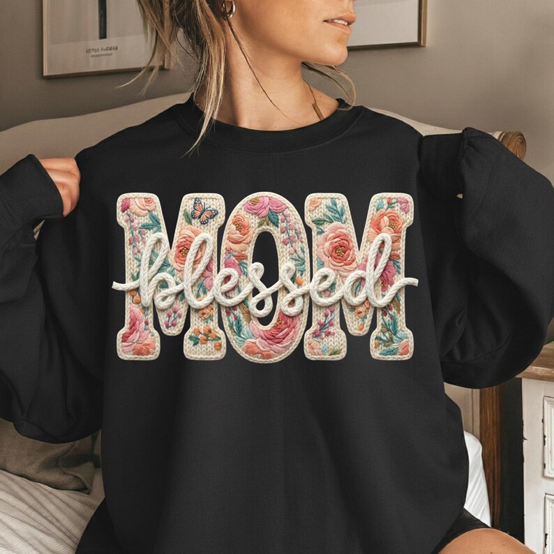 May include: Black sweatshirt with the words "MOM blessed" in a floral design. The letters are filled with pink, peach, and green flowers, and a small butterfly is visible. The word "blessed" is in a cursive font.