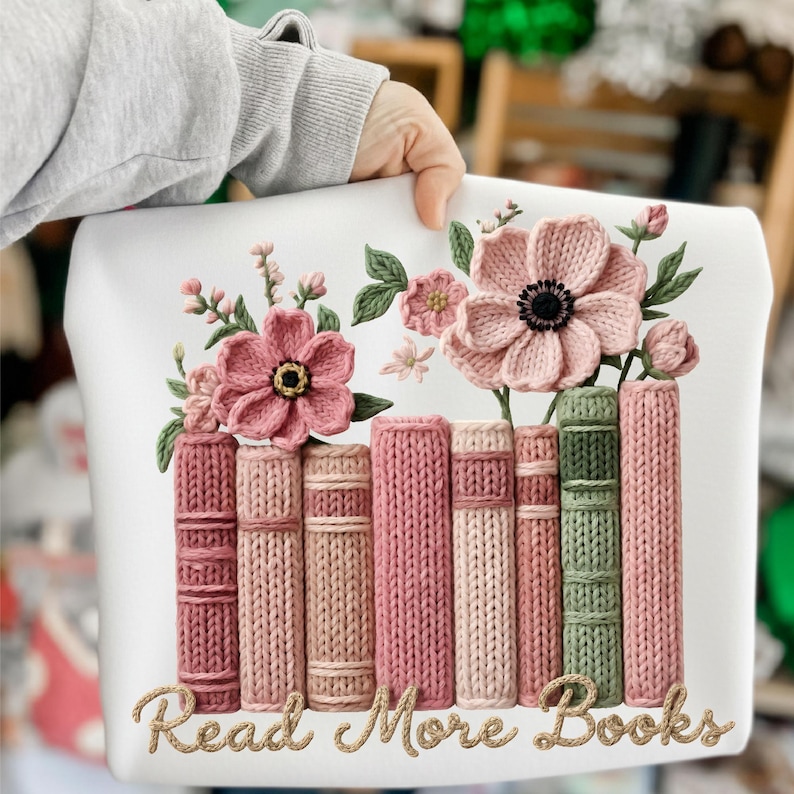 May include: White decorative pillow featuring a floral and book design. The design includes pink flowers, green leaves, and a row of books in shades of pink and green. The text "Read More Books" is written in a cursive font.