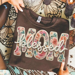 May include: A brown sweatshirt with the word "MOM" and the word "Blessed" embroidered on the front. The letters are filled with floral designs in shades of pink, orange, and green, with a small butterfly accent.