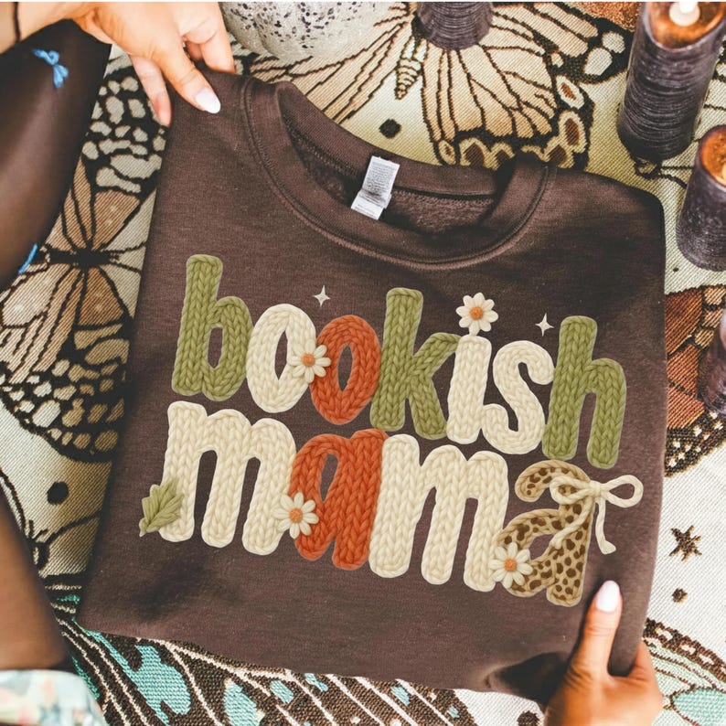 May include: A brown sweatshirt with the words "bookish mama" in a textured, colorful design. The letters are in shades of green, orange, and cream, with floral and leopard print accents. The sweatshirt is held by a person.